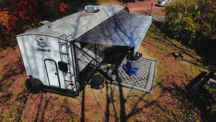 Aerial view of an RV rental setup at an autumn campsite, with an extended awning, outdoor rug, and chairs.. Jayco White Hawk 2022