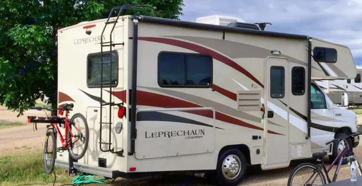 A beige Coachmen Leprechaun Class C **rv rental** is parked at a campsite with bicycles on its rear rack and in the foreground.. Forest River Coachmen Leprechaun 2017
