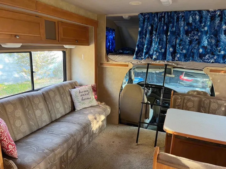 Spacious RV rental interior featuring a comfortable sofa, overhead bunk with blue curtains, and a bright window view.. Winnebago Access 2008