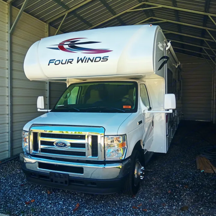 A white Ford Four Winds Class C rv rental is parked on gravel under a metal shelter.. Thor Four Winds 2022