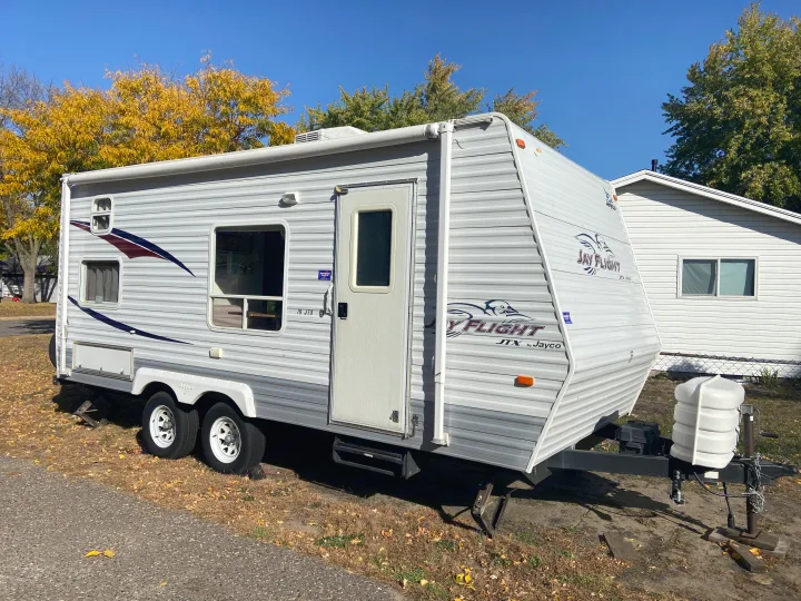 White Jayco Jay Flight JTX travel trailer RV rental, featuring two axles and propane tanks, parked outdoors on a sunny fall day.. Jayco Jay Flight 2007