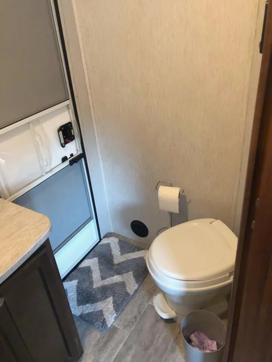 RV rental bathroom with toilet, TP holder, and a gray chevron mat. Screen door to the left, showing compact design.. Heartland RVs Mallard 2019