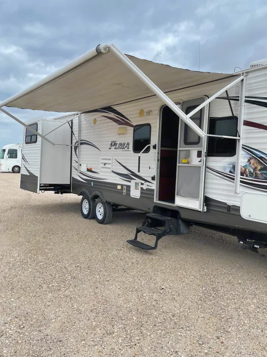 A white Puma RV rental with its awning and slide-out extended, parked on gravel. Another RV is in the background.. Palomino Puma 2015