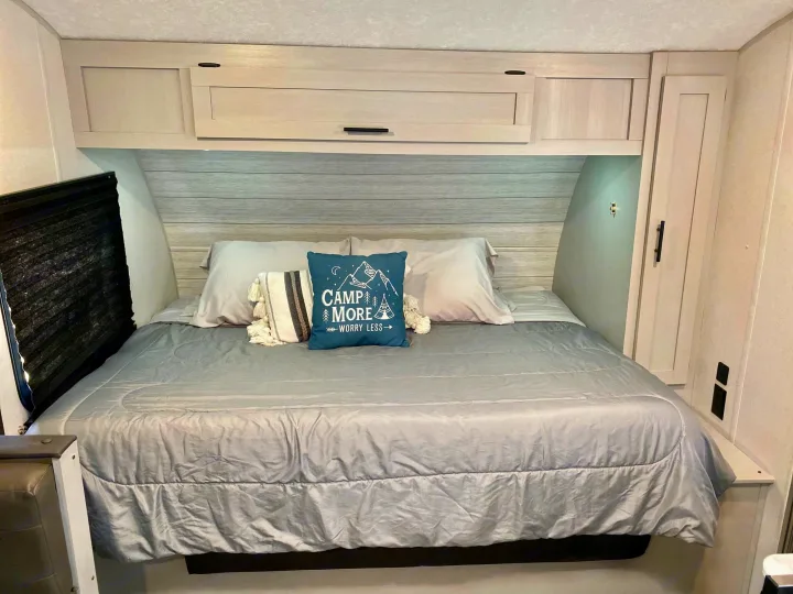 An RV rental bedroom with a comfy grey bed, overhead cabinets, and "CAMP MORE WORRY LESS" pillow offers a relaxing retreat.. Forest River Wildwood 2021