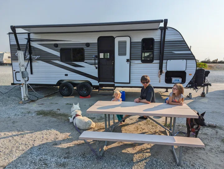 Family and dogs relax by their RV rental at a sunny campsite, enjoying a picnic by their travel trailer.. Highland Ridge RV Open Range 2022