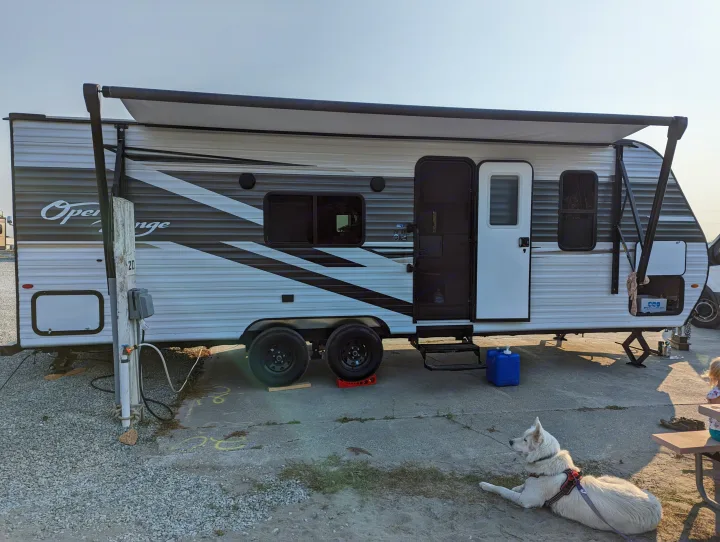 Sunny RV rental travel trailer at a campsite, awning extended, with a white dog relaxing nearby.. Highland Ridge RV Open Range 2022