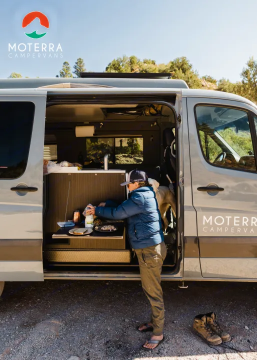 Enjoying van life with an RV rental. A person prepares food outside a Moterra Campervan, ready for outdoor adventure.. Mercedes-Benz Sprinter 2021
