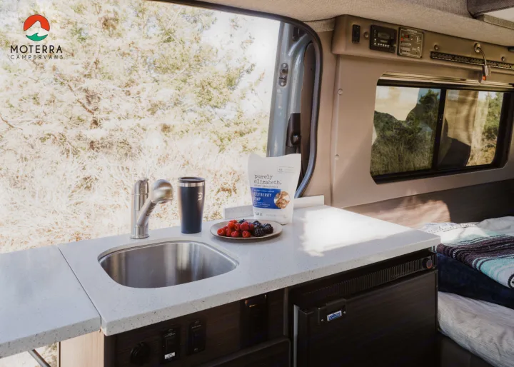 Inside an rv rental: a modern campervan kitchen with sink, fresh fruit, and fridge, ready for your outdoor adventures.. Mercedes-Benz Sprinter 2021