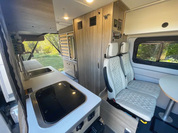 Inside a modern RV rental: kitchenette, dinette with sleek white seats, and versatile sleeping area in a bright camper van.. Winnebago Solis PX 2023