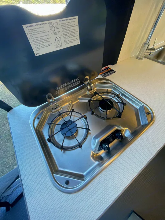 Compact RV rental kitchen featuring a two-burner stainless steel cooktop, open lid with lighting instructions, and a sink.. Winnebago Solis PX 2023
