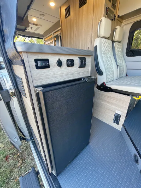 Inside an RV rental: a compact refrigerator, control panel, and comfortable passenger seating in a camper van.. Winnebago Solis PX 2023