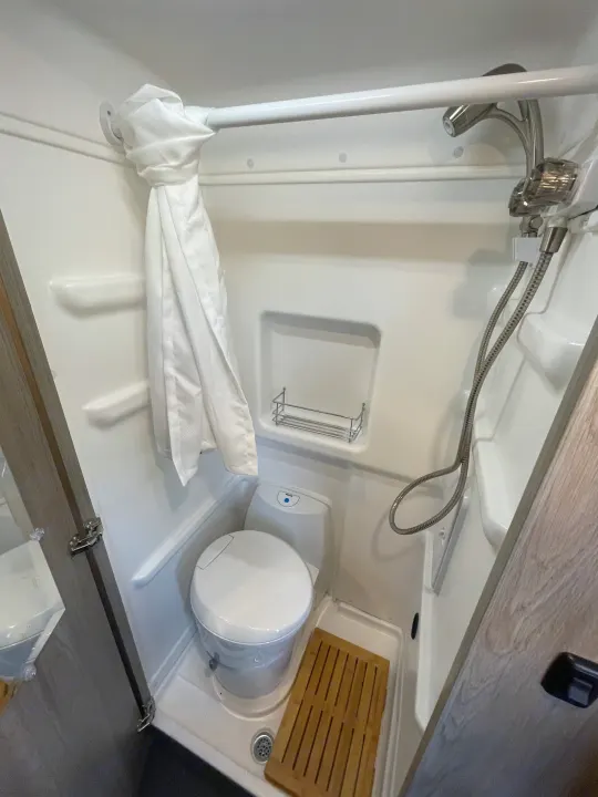 RV rental bathroom with a compact design, featuring a toilet, shower, curtain, shelf, and wooden floor mat.. Winnebago Solis PX 2023