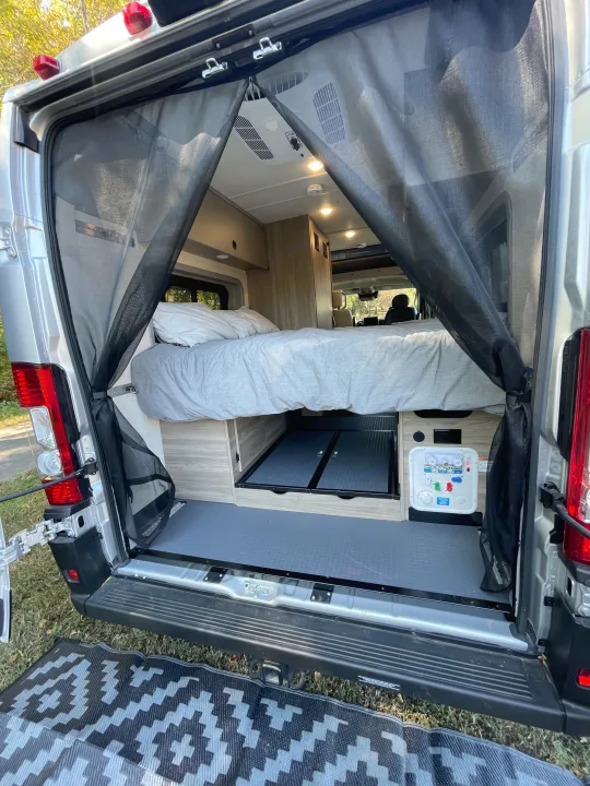 RV rental: Rear view of a camper van, showcasing its bed, interior storage, and a magnetic screen door. Outdoor mat visible.. Winnebago Solis PX 2023