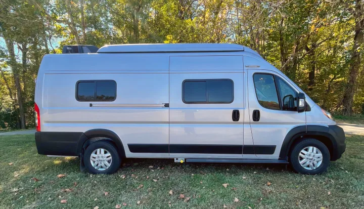 Side view of a silver Class B RV rental camper van, parked on grass in front of green trees.. Winnebago Solis PX 2023