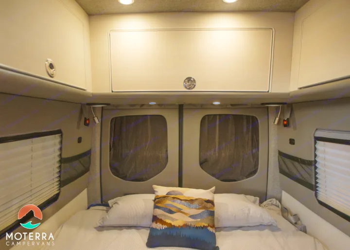 Interior of an RV rental: a cozy bed with pillows, overhead storage cabinets, and ambient lighting. Window blinds visible.. Mercedes-Benz Sprinter 2020