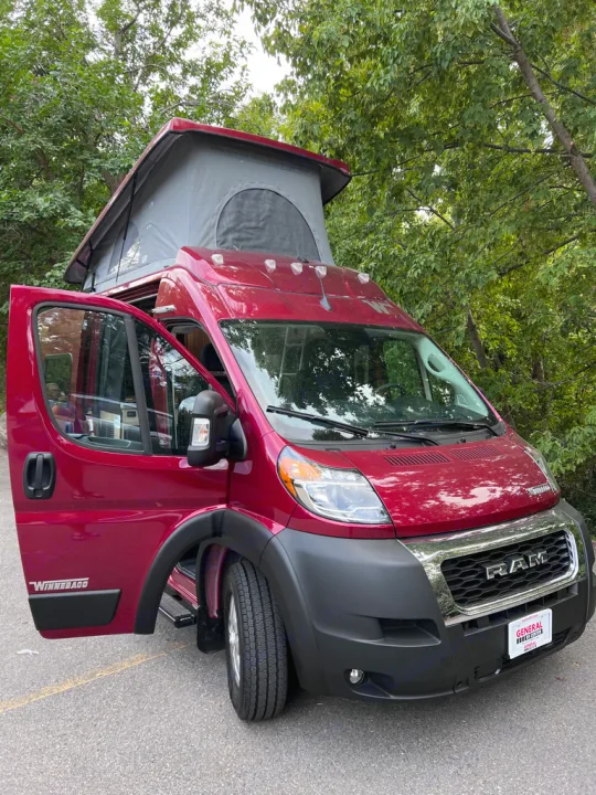 This red Winnebago camper van, an RV rental, features an extended pop-top and open side door, parked among trees.. Winnebago Solis 59PX 2023