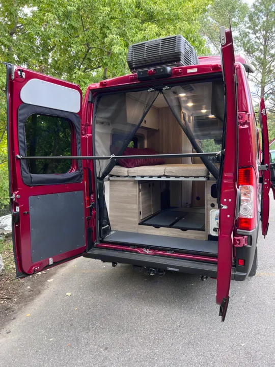 Inside an RV rental, the open rear of this red camper van shows a raised bed, mesh screen, and ample storage.. Winnebago Solis 59PX 2023