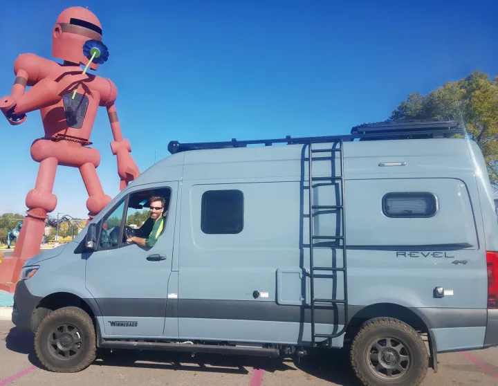 Smiling man in an RV rental (Winnebago Revel 4x4 camper van) next to a giant red robot holding a flower.. Winnebago Revel 2020
