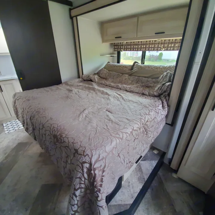 Comfortable RV rental bedroom featuring a patterned bed, light wood cabinets with dark handles, and a window with blinds.. Forest River Sunseeker 2020