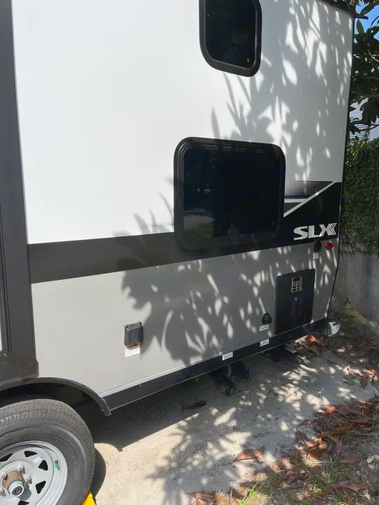 Side of an SLX rv rental travel trailer, with utility hookups & wheel. Parked under dappled tree shadows.. Jayco 184bs Bunk House Jay Flight SLX 2022