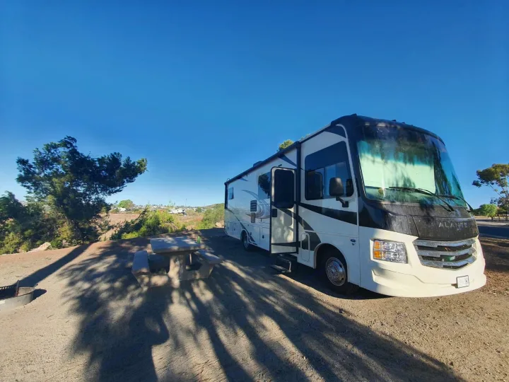 Alante RV rental parked at a sunlit campsite, featuring a concrete picnic table and fire pit.. Jayco Alante 2022