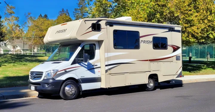 Tan and white Mercedes-Benz Sprinter RV rental parked on asphalt next to a tennis court, ready for travel adventures.. Forest River Coachmen Prism 2018