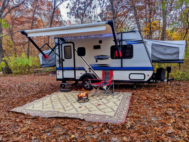 An RV rental travel trailer set up with an awning, outdoor mat, warm campfire, and chair amidst vibrant autumn leaves.. Jayco Jay Feather 2018