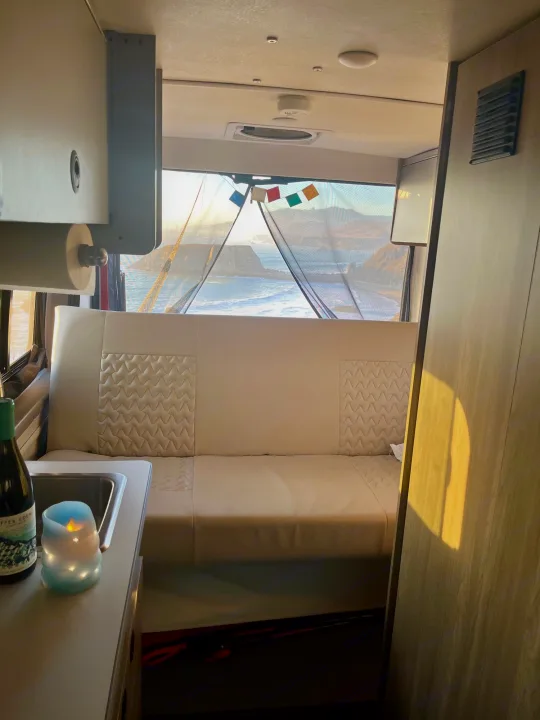 Cozy RV rental interior: a comfy bench and small sink face a large window offering stunning ocean and coastal rock views at sunset.. Winnebago Solis 2021