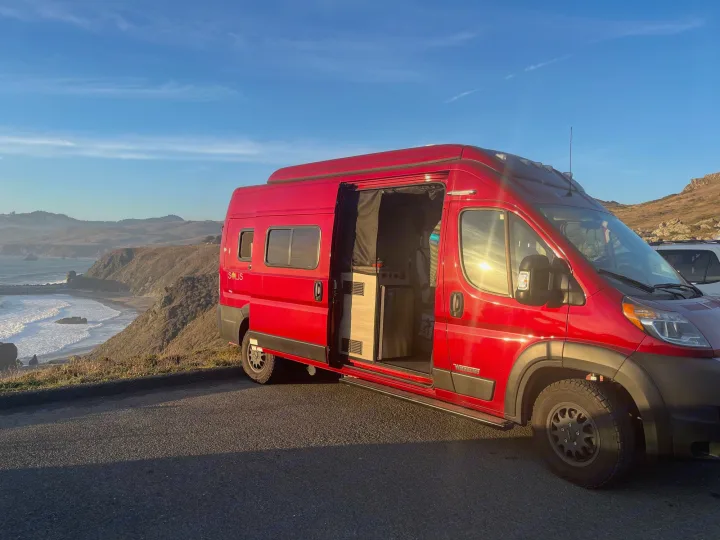 A red Winnebago Solis rv rental camper van on a scenic coastal road trip overlook. Door open, interior visible.. Winnebago Solis 2021