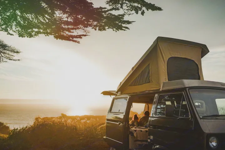 This RV rental offers stunning ocean sunset views. A dark pop-top camper van is parked, with two people relaxing inside.. Volkswagen Vanagon 1985
