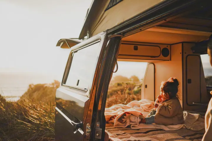 Two kids cozy up inside an RV rental camper van with its pop-top open, parked on a sunny, grassy coast.. Volkswagen Vanagon 1985