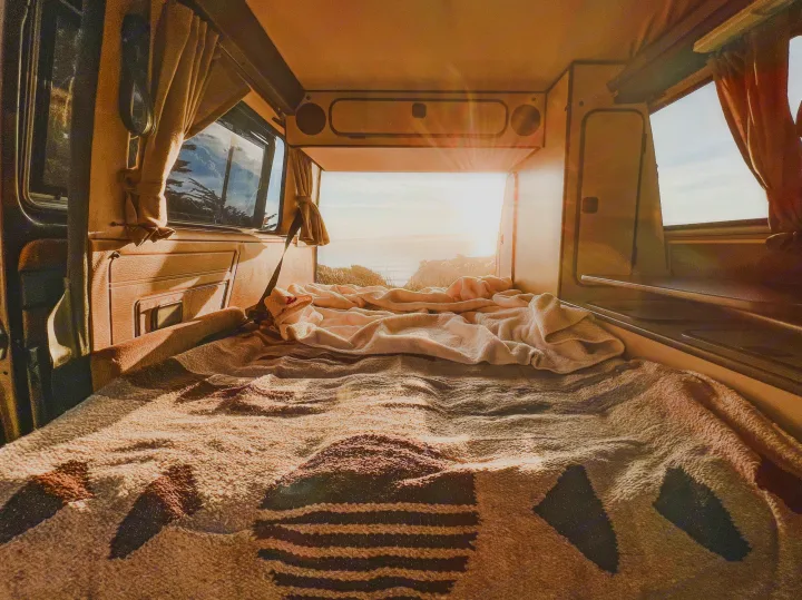 Cozy RV rental interior shows a made bed, patterned blankets, and a bright ocean sunset view from the rear.. Volkswagen Vanagon 1985