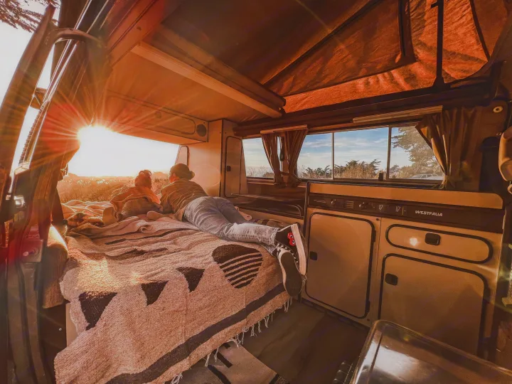 A person relaxing on a bed in an RV rental camper van, watching a vibrant sunset through the rear window. Cozy van life.. Volkswagen Vanagon 1985