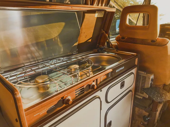 Cozy Westfalia rv rental interior showing a vintage kitchenette with stove and sink bathed in warm sunlight, perfect for adventures.. Volkswagen Vanagon 1985