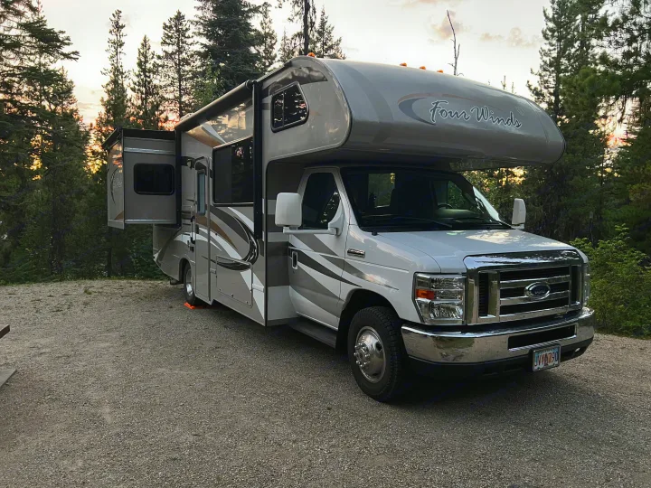 A Class C Four Winds RV rental, with its slide-out extended, is parked on a gravel campsite at sunset among tall trees.. Thor Four Winds 2014