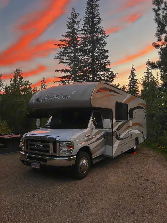 An RV rental parked amidst tall trees in a tranquil forest, bathed in the warm glow of a pink and orange sunset.. Thor Four Winds 2014