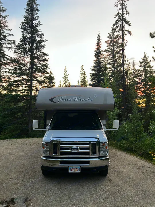 White Four Winds **rv rental** parked on a gravel road among tall pine trees, ready for a forest adventure.. Thor Four Winds 2014