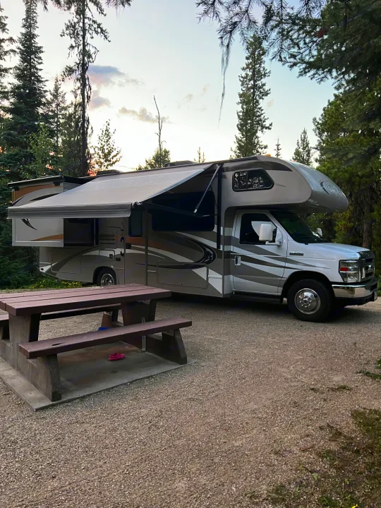 Class C RV rental at a gravel campsite among tall trees, with awning extended and a picnic table ready for use.. Thor Four Winds 2014