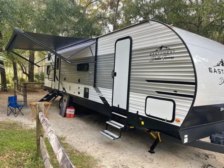 A sleek East West Della Terra travel trailer, available for rv rental, is parked at a shaded campsite with its awning extended.. Della Terra 312H Della Terra 2023