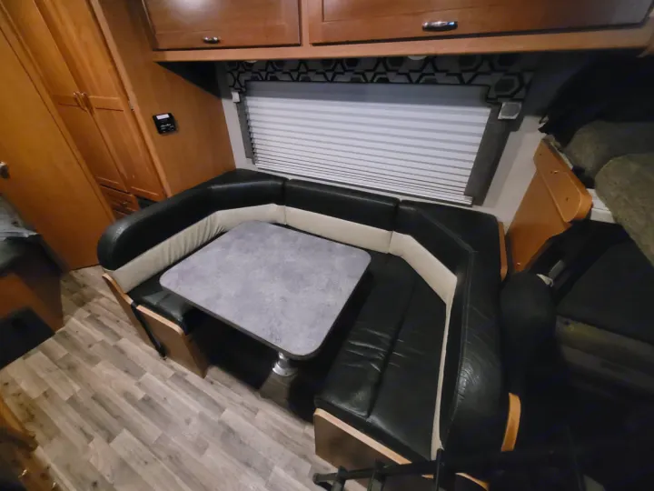 RV rental interior features a U-shaped dinette with black/white booth seating, grey table, wood cabinets, and a window blind.. Winnebago Minnie Winnie 2018