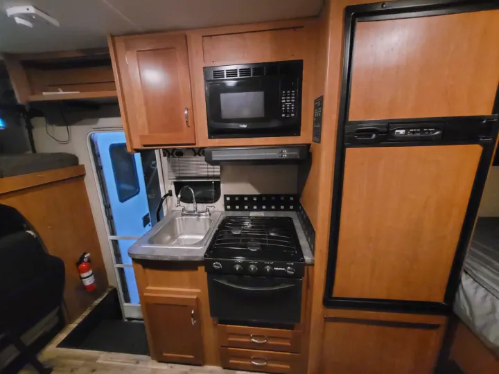 Inside this RV rental: a compact kitchen with stove, microwave, sink, and wood-paneled fridge. Fire extinguisher near exit.. Winnebago Minnie Winnie 2018