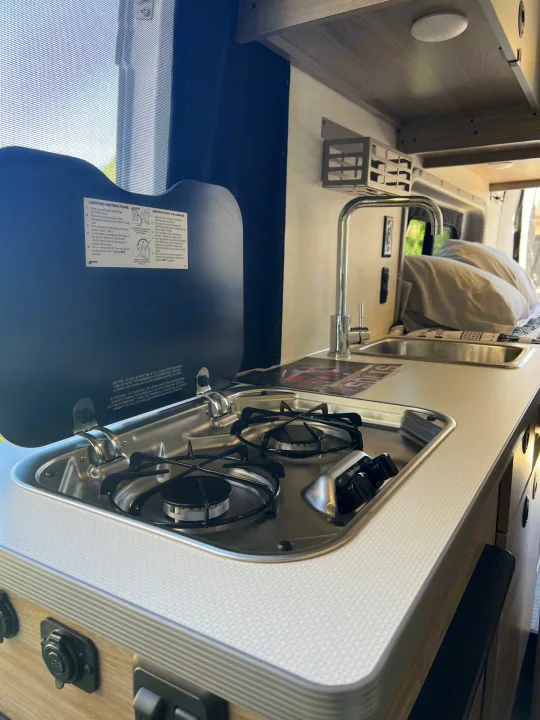 RV rental camper van kitchen features a two-burner gas stove with lid instructions and a sink for travel cooking.. Winnebago Solis 2023