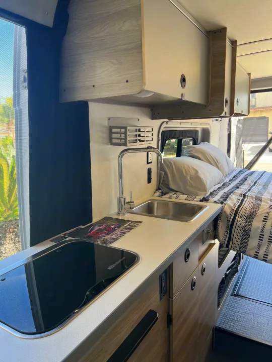 This RV rental's interior highlights a compact kitchenette with sink, stovetop, and comfortable bed, plus overhead storage.. Winnebago Solis 2023