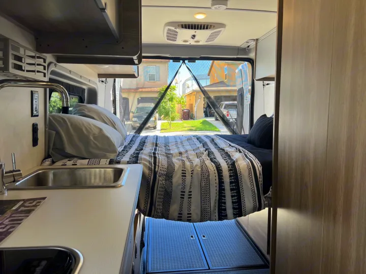RV rental interior: cozy bed, compact kitchen sink, and open rear doors reveal a sunny residential street. Mobile living comfort.. Winnebago Solis 2023
