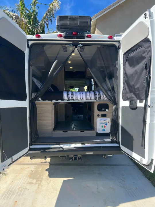 Step inside this cozy rv rental! Rear view shows open campervan doors with bug screens, bed, storage, and utility panel.. Winnebago Solis 2023