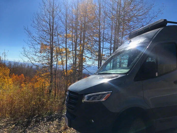 Gray camper van RV rental parked amidst vibrant fall foliage and distant mountains under a sunny blue sky.. Winnebago Revel 2023