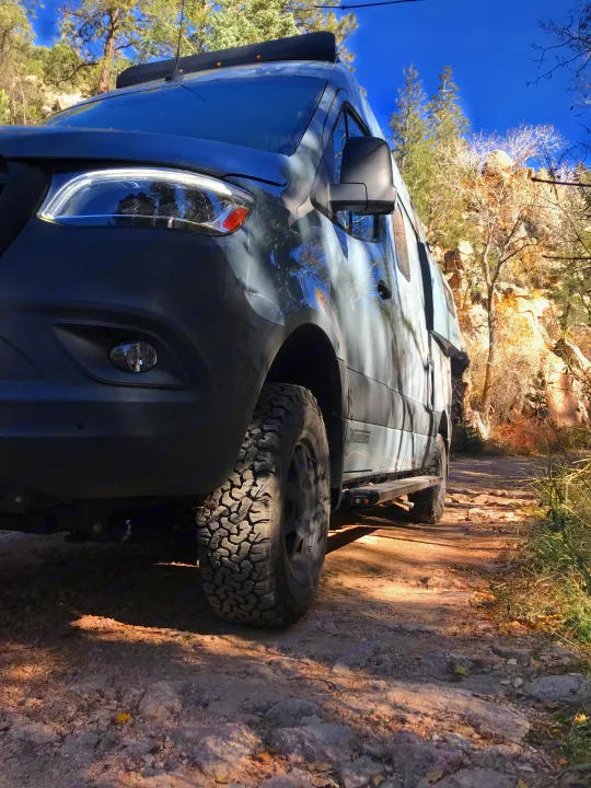 An off-road capable RV rental, possibly a Winnebago, on a rocky dirt trail through a scenic, sunny, forested area.. Winnebago Revel 2023