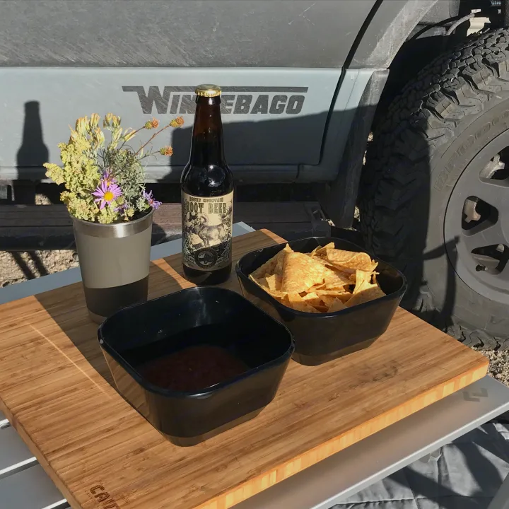 An RV rental provides chips, salsa, root beer, and wildflowers on a portable table next to a Winnebago, ideal for outdoor relaxing.. Winnebago Revel 2023