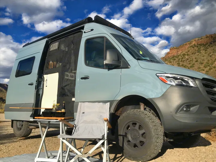 Grey camper van RV rental parked in a scenic outdoor setting, side door open, with an outdoor table and chair setup.. Winnebago Revel 2023