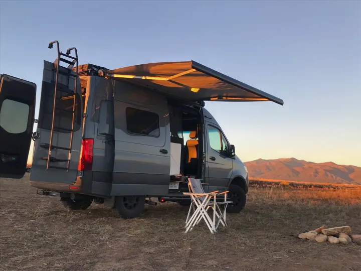 Ideal **rv rental** campsite: gray camper van, open doors, extended awning, and a camp chair in a field with mountains at sunset.. Winnebago Revel 2023
